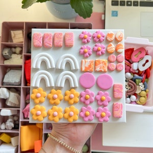 May include: A white board with a variety of colorful polymer clay earrings. The earrings are in the shapes of flowers, arches, and swirls. The board is divided into sections with each section containing a different type of earring.