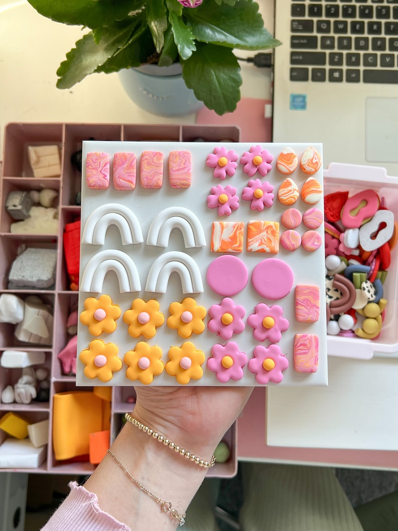 May include: A white board with a variety of colorful polymer clay earrings. The earrings are in the shapes of flowers, arches, and swirls. The board is divided into sections with each section containing a different type of earring.