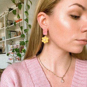 May include: A person wearing a pink sweater and a gold chain necklace with a teardrop pendant. They are wearing a pair of yellow and pink flower-shaped earrings.