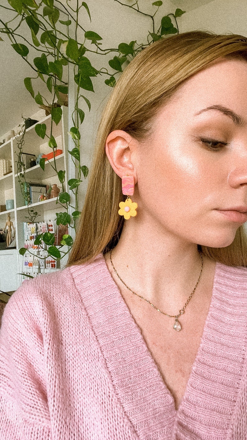 May include: A person wearing a pink sweater and a gold chain necklace with a teardrop pendant. They are wearing a pair of yellow and pink flower-shaped earrings.