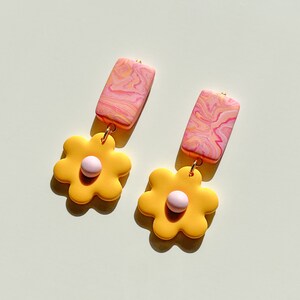 May include: A pair of yellow flower-shaped earrings with pink centers and pink and white marbled rectangular dangles.