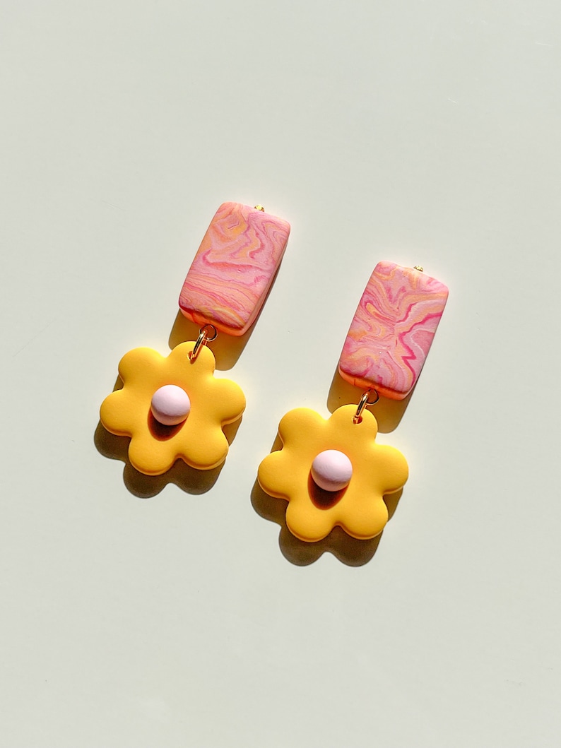 May include: A pair of yellow flower-shaped earrings with pink centers and pink and white marbled rectangular dangles.