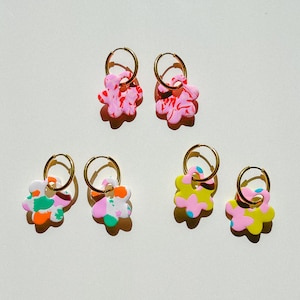 Op de afbeelding: Zes paar kleurrijke bloemvormige oorbellen met goudkleurige ringen. De oorbellen hebben abstracte patronen in roze, geel, groen, oranje en blauw. Ze worden gepresenteerd op een witte ondergrond.