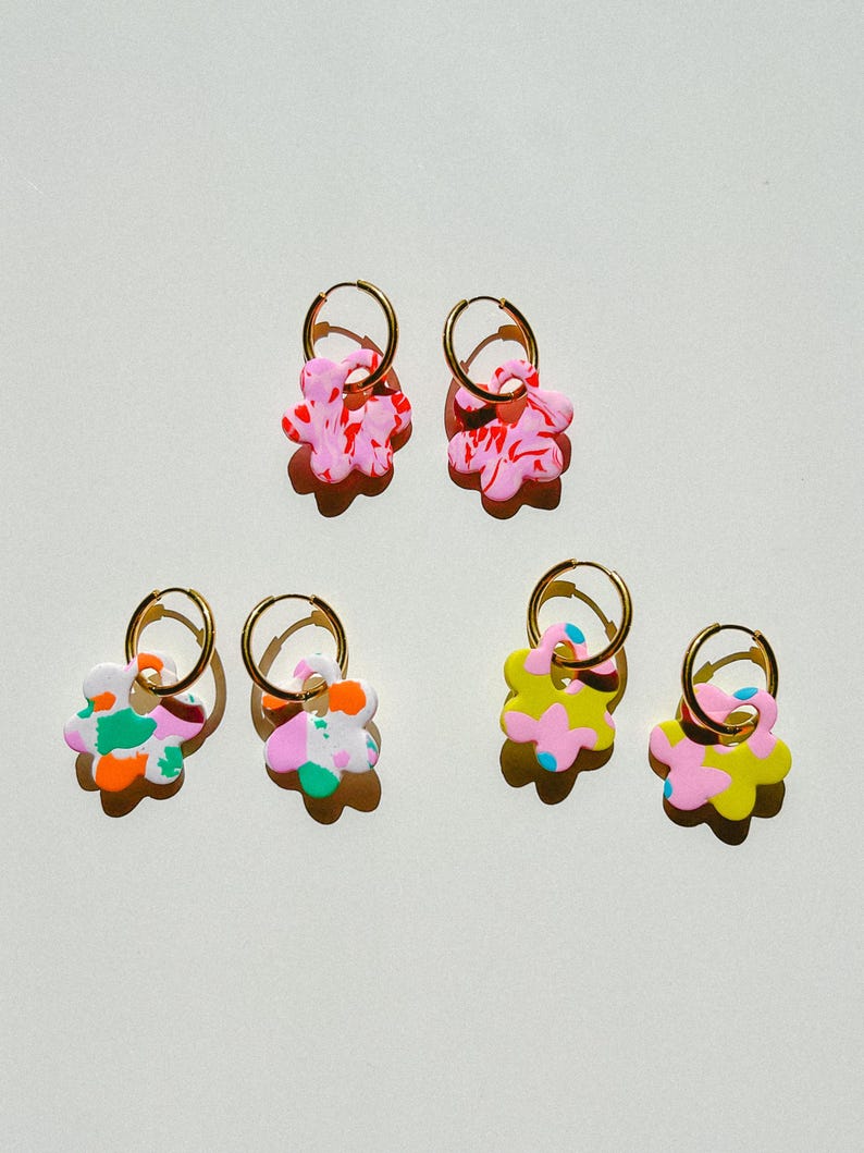 Op de afbeelding: Zes paar kleurrijke bloemvormige oorbellen met goudkleurige ringen. De oorbellen hebben abstracte patronen in roze, geel, groen, oranje en blauw. Ze worden gepresenteerd op een witte ondergrond.
