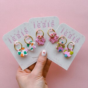 Op de afbeelding: Een hand houdt meerdere paren bloemvormige oorbellen op kaarten vast. De oorbellen hebben gouden hoepels en kleurrijke bloemmotieven in roze, groen, oranje en blauw. De kaarten hebben de tekst "studio iebis" en een websiteadres.