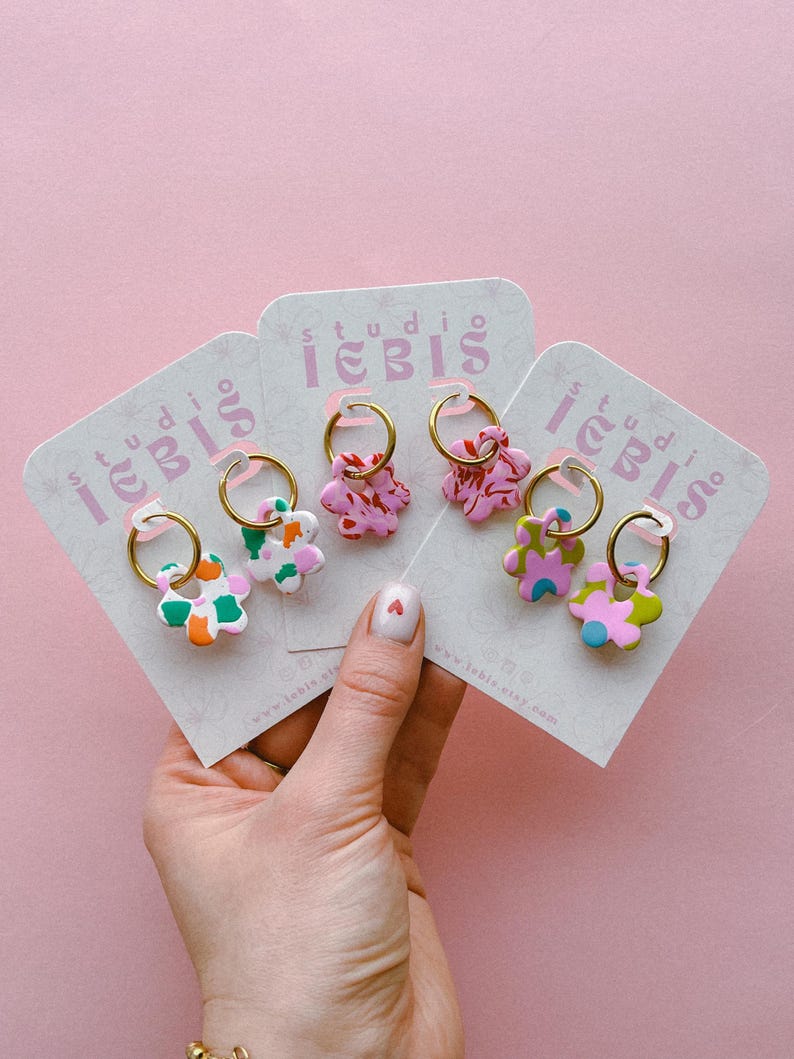 Op de afbeelding: Een hand houdt meerdere paren bloemvormige oorbellen op kaarten vast. De oorbellen hebben gouden hoepels en kleurrijke bloemmotieven in roze, groen, oranje en blauw. De kaarten hebben de tekst "studio iebis" en een websiteadres.