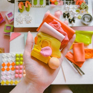 May include: A hand holding a stack of colorful polymer clay, including yellow, pink, orange, and red. The clay is arranged in a stack, with the yellow clay on top. There is a small pink ball of clay next to the stack.