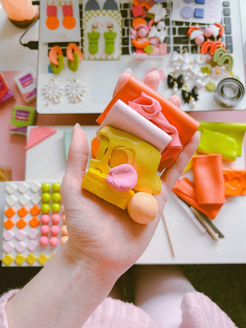 May include: A hand holding a stack of colorful polymer clay, including yellow, pink, orange, and red. The clay is arranged in a stack, with the yellow clay on top. There is a small pink ball of clay next to the stack.