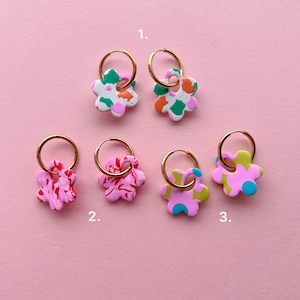 Op de afbeelding: Een verzameling bloemvormige oorbellen met gouden creolen. De oorbellen hebben verschillende kleurencombinaties, waaronder wit, groen, oranje, roze en blauw. De oorbellen worden gepresenteerd tegen een roze achtergrond.