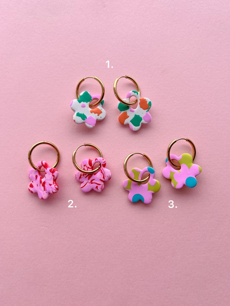 Op de afbeelding: Een verzameling bloemvormige oorbellen met gouden creolen. De oorbellen hebben verschillende kleurencombinaties, waaronder wit, groen, oranje, roze en blauw. De oorbellen worden gepresenteerd tegen een roze achtergrond.