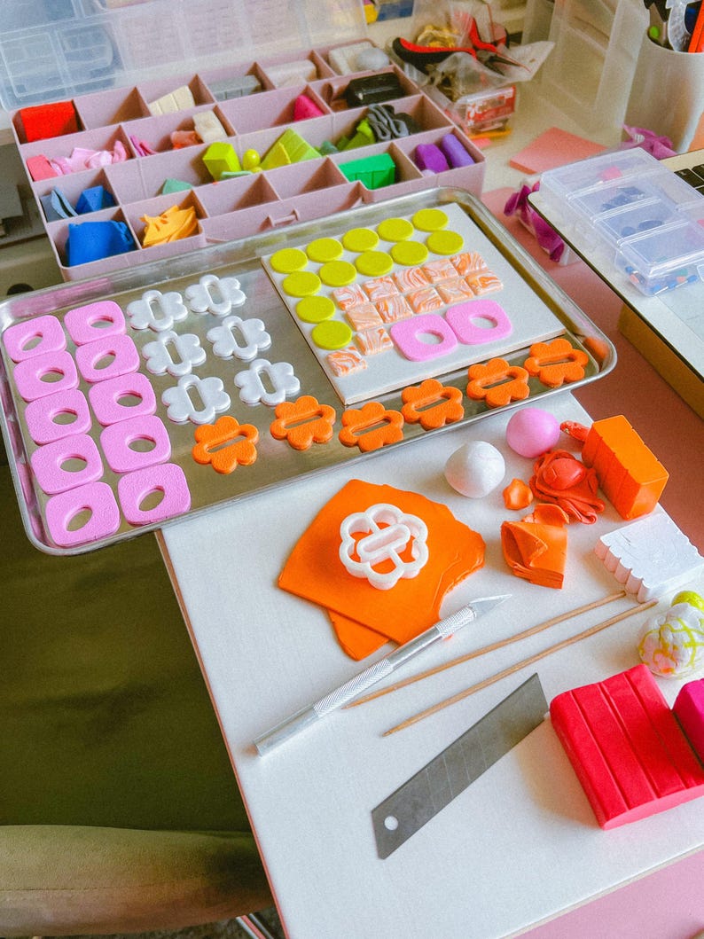 Op de afbeelding: Een werkplek met polymeerklei en gereedschap. Roze, witte en oranje kleivormen zijn gerangschikt op een metalen dienblad. Een roze organizer bevat diverse kleuropties. Gereedschap omvat een hobbymes, houten spiesjes en een metalen mes.