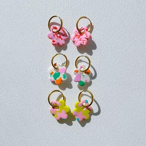 Op de afbeelding: Zes paar bloemvormige oorbellen met gouden hoepels. De oorbellen hebben roze, witte, groene, oranje en gele ontwerpen. De oorbellen zijn symmetrisch gerangschikt tegen een witte achtergrond.