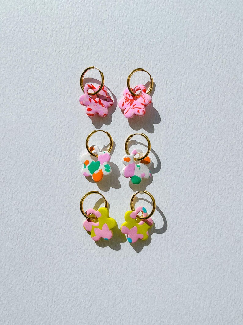 Op de afbeelding: Zes paar bloemvormige oorbellen met gouden hoepels. De oorbellen hebben roze, witte, groene, oranje en gele ontwerpen. De oorbellen zijn symmetrisch gerangschikt tegen een witte achtergrond.