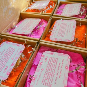 Op de afbeelding: Open kartonnen dozen gevuld met roze en oranje vloeipapier en zilveren snippers. Elke doos bevat een roze en wit bloemig patroon kaart met tekst. De dozen zijn gerangschikt in een raster.