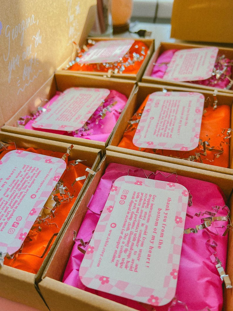 Op de afbeelding: Open kartonnen dozen gevuld met roze en oranje vloeipapier en zilveren snippers. Elke doos bevat een roze en wit bloemig patroon kaart met tekst. De dozen zijn gerangschikt in een raster.
