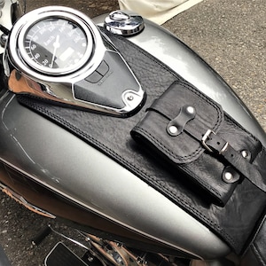 Può includere: Una borsa da serbatoio per moto in pelle nera con chiusura a fibbia. La borsa è fissata a un serbatoio di benzina per moto argentato.