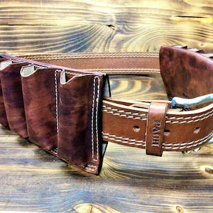 Pode incluir: Um cinto de couro marrom com uma fivela de prata e vários laços para segurar ferramentas. O cinto é costurado com fio branco e tem a palavra "PACH" gravada na fivela.