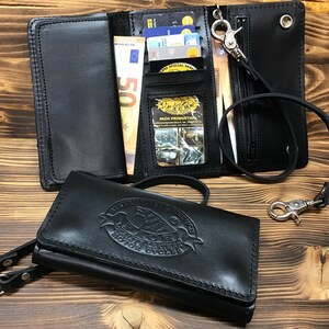 Può includere: Portafoglio in pelle nera con una catena e una chiusura a scatto. Il portafoglio ha più slot per carte e un portafoglio. Il portafoglio ha un logo di aquila in rilievo con il testo "Leather Motorcycle Gear".