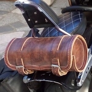 Può includere: Una borsa da sella per moto in pelle marrone con un design cucito e fibbie. La borsa da sella è fissata alla parte posteriore di una moto.
