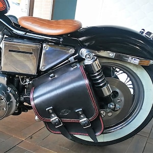Peut inclure: Sacoche de moto en cuir noir avec des coutures et des boucles rouges, fixée à une moto noire avec un pneu à flancs blancs.