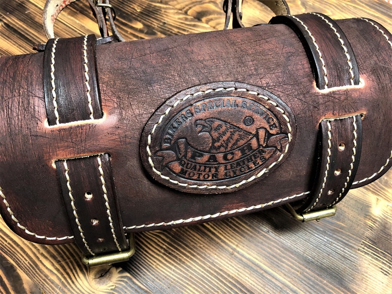 May include: Brown leather motorcycle saddlebag with white stitching and a brass buckle. The bag has a circular logo with the text "Bikers Special Service" and "Paci Quality Leather Motorcycles".