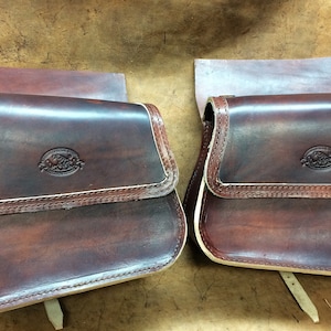 Può includere: Due bisacce in pelle marrone con chiusura a patta e cuciture decorative. Ogni borsa presenta un piccolo stemma in rilievo e un bordo colorato chiaro. Le borse hanno una forma rettangolare e sono progettate per una sella.