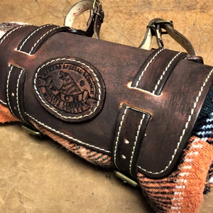 May include: A brown leather blanket roll with a strap and buckle. The roll has a stitched design and a leather patch with the text "Riders Special Service Quality Leather Motor Cycles" and a logo of a bird.