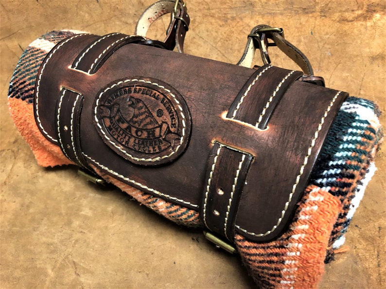 May include: A brown leather blanket roll with a strap and buckle. The roll has a stitched design and a leather patch with the text "Riders Special Service Quality Leather Motor Cycles" and a logo of a bird.
