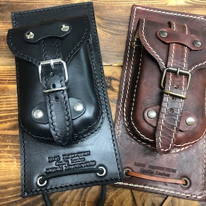 May include: Two leather phone holsters with a strap and buckle closure. The holster on the left is black and the holster on the right is brown. Both holsters have white stitching and the text "BACK PRODUCTION Quality Leather" is embossed on the bottom.