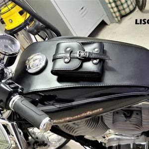 May include: Black leather motorcycle tank bag with a buckle closure. The bag is attached to a black motorcycle petrol tank with a chrome Harley Davidson emblem.