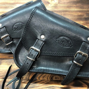 May include: Two black leather motorcycle saddlebags with buckles and stitching. Each bag has a logo embossed on the front.