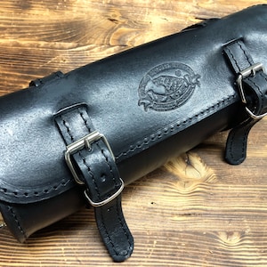 Può includere: Sella da moto in pelle nera con un design cucito e fibbie argentate. La borsa ha un logo in rilievo con il testo "The Garage".