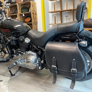 Può includere: Una motocicletta Harley-Davidson nera con serbatoio lucido, sella in pelle nera con schienale e una grande borsa laterale in pelle nera con due fibbie argentate sul lato. I componenti del motore sono di colore argento e nero.