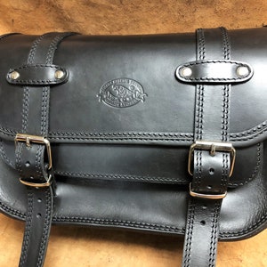 Può includere: Sella da moto in pelle nera con due fibbie e un design cucito. La borsa ha un logo del marchio in rilievo sul davanti.
