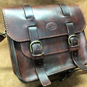 May include: A brown leather messenger bag with two adjustable straps and buckles. The bag has a flap with a stitched design and a metal logo that says "PAUL".