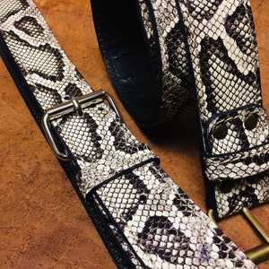 Può includere: Una cintura in pelle marrone con un motivo a pelle di serpente e una fibbia argentata. La cintura è piegata a metà e la fibbia è visibile.