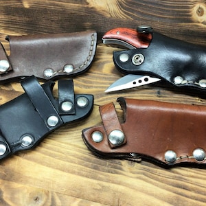 Könnte beinhalten: Vier Leder-Messerscheiden in Braun- und Schwarztönen. Eine Scheide enthält ein Messer mit Holzgriff. Jede Scheide ist mit Metallnieten und Nähten befestigt. Präsentiert auf einer Holzoberfläche.