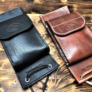 Op de afbeelding: Twee leren telefoonhoesjes met een klep sluiting en een band. Het hoesje links is zwart en het hoesje rechts is bruin. Beide hoesjes hebben de tekst "BACH PRODUCTION Quality Leather" op de achterkant.