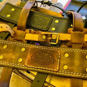 May include: Two leather tool belts with brass rivets and stitching. The top belt is black with a brown leather logo that reads "The Leather Stocking Co.  Genuine Leather". The bottom belt is brown with a brown leather logo that reads "The Leather Stocking Co.  Genuine Leather".