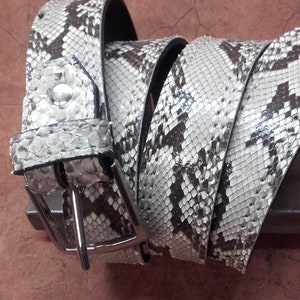 以下が含まれることがあります： 白と黒のヘビ革のベルトに銀色のバックルが付いている。ベルトは巻かれており、バックルが見える。