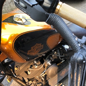 May include: Close-up of a custom motorcycle with a gold and black gas tank. The tank has a Harley-Davidson logo and a black leather seat. The motorcycle has a black leather handlebar with a gold throttle.