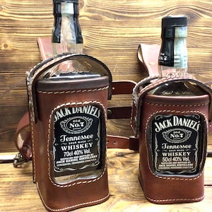 Könnte beinhalten: Zwei braune Leder-Jack-Daniel's-Whiskey-Flaschenhalter mit Riemen. Die Flaschenhalter sind aus braunem Leder gefertigt und haben ein transparentes Fenster, um das Flaschenetikett zu zeigen. Die Flaschenhalter sind beide mit "Jack Daniel's Old No. 7 Tennessee Whiskey" beschriftet.
