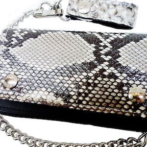 Könnte beinhalten: Eine silberne Kettengeldbörse mit einer schwarzen Lederklappe und einem Schlangenhautmuster. Die Geldbörse hat zwei silberne Druckknöpfe und eine silberne Kette.