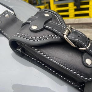 May include: Black leather motorcycle accessory with white stitching and silver hardware. The accessory has a buckle closure and is designed to attach to a motorcycle. The item is shown on a grey surface.