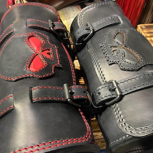 Può includere: Due protezioni per braccia in pelle nera con cuciture rosse e ritagli decorativi. Uno presenta un design a teschio, l'altro un giglio. Ognuno ha due fibbie regolabili per una vestibilità sicura.