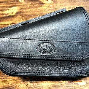May include: Black leather motorcycle saddlebag with a stitched flap and a logo that reads "Genuine Leather Goods, Handcrafted in the USA".
