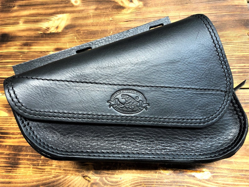 May include: Black leather motorcycle saddlebag with a stitched flap and a logo that reads "Genuine Leather Goods, Handcrafted in the USA".