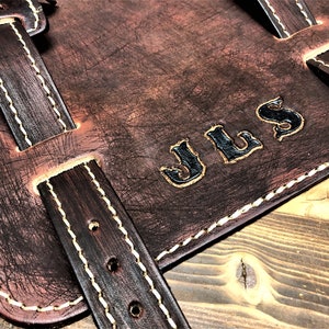May include: Brown leather motorcycle saddlebag with white stitching and a black leather strap. The bag has the initials "JLS" embossed in black on the front.