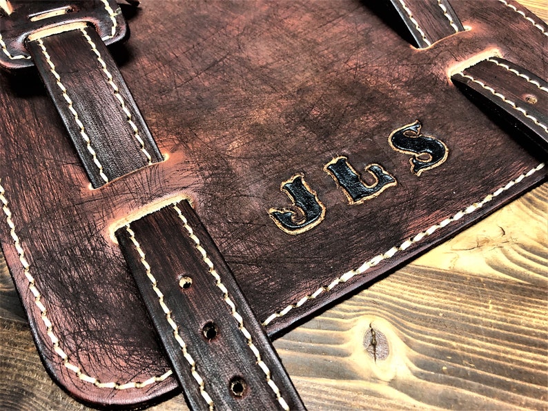 May include: Brown leather motorcycle saddlebag with white stitching and a black leather strap. The bag has the initials "JLS" embossed in black on the front.