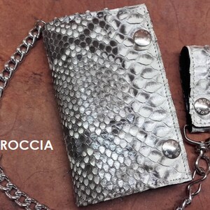 Può includere: Un portafoglio in pelle di serpente argentato con una catena e un portachiavi abbinato. Il portafoglio ha la scritta "ROCCIA" stampata su di esso.